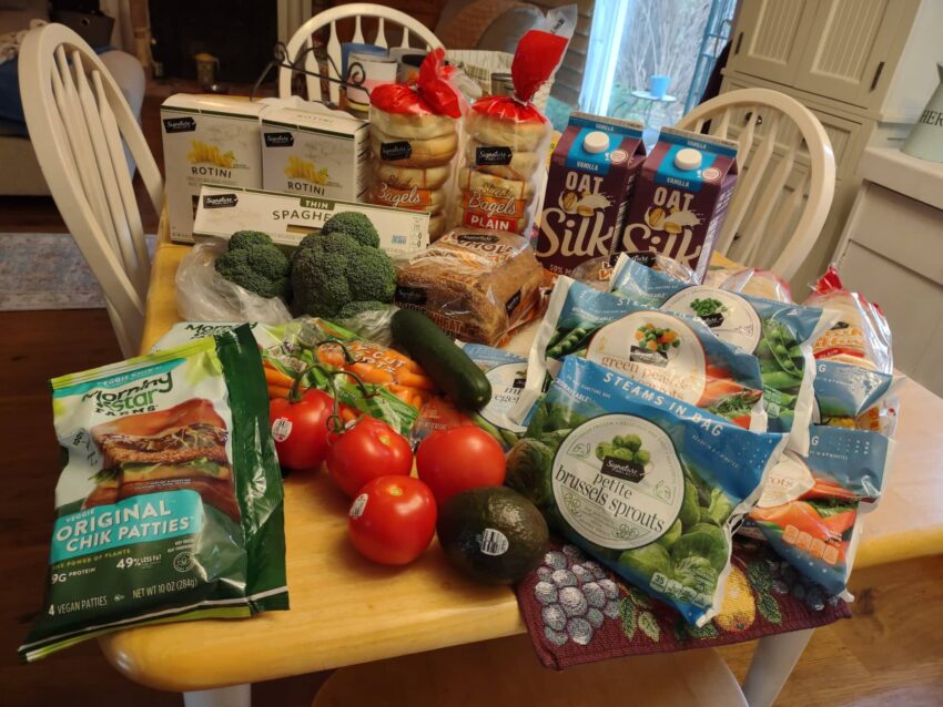 My Safeway groceries on dining table includes Silk oatmilk, packs of frozen veggies, Mornigstar Farms chik'n burgers, avocado, tomato, broccoli, bagels, English muffins, sandwich bread, cucumber, baby carrots, boxes of pasta