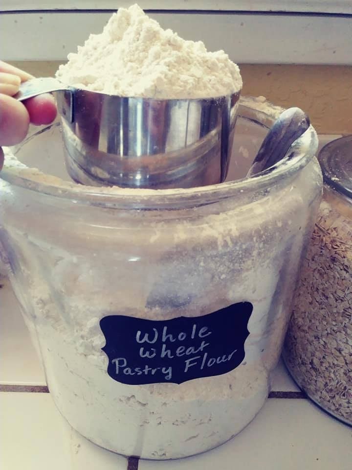 glass canister that says whole wheat pastry flour with me scooping out a cup with a metal cup